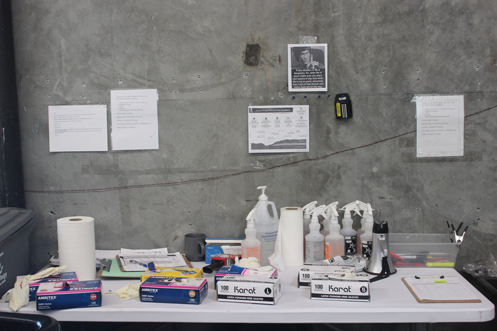 Sanitation supplies including clothes, bottles of disinfectant, and paper towels are sprawled across a white plastic table, along with administrative supplies including clip boards and highlighters.